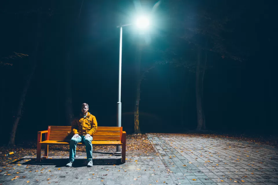 Man sitting on wooden bench under tall streetlight at night.